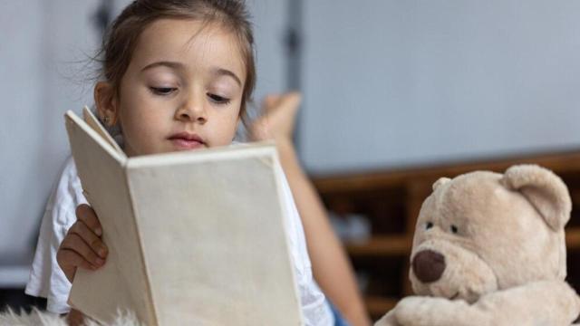 Una niña lee junto a un oso de peluche