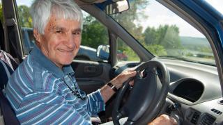 Un hombre mayor sonríe mientras está al volante de un coche, en un entorno natural y de día.