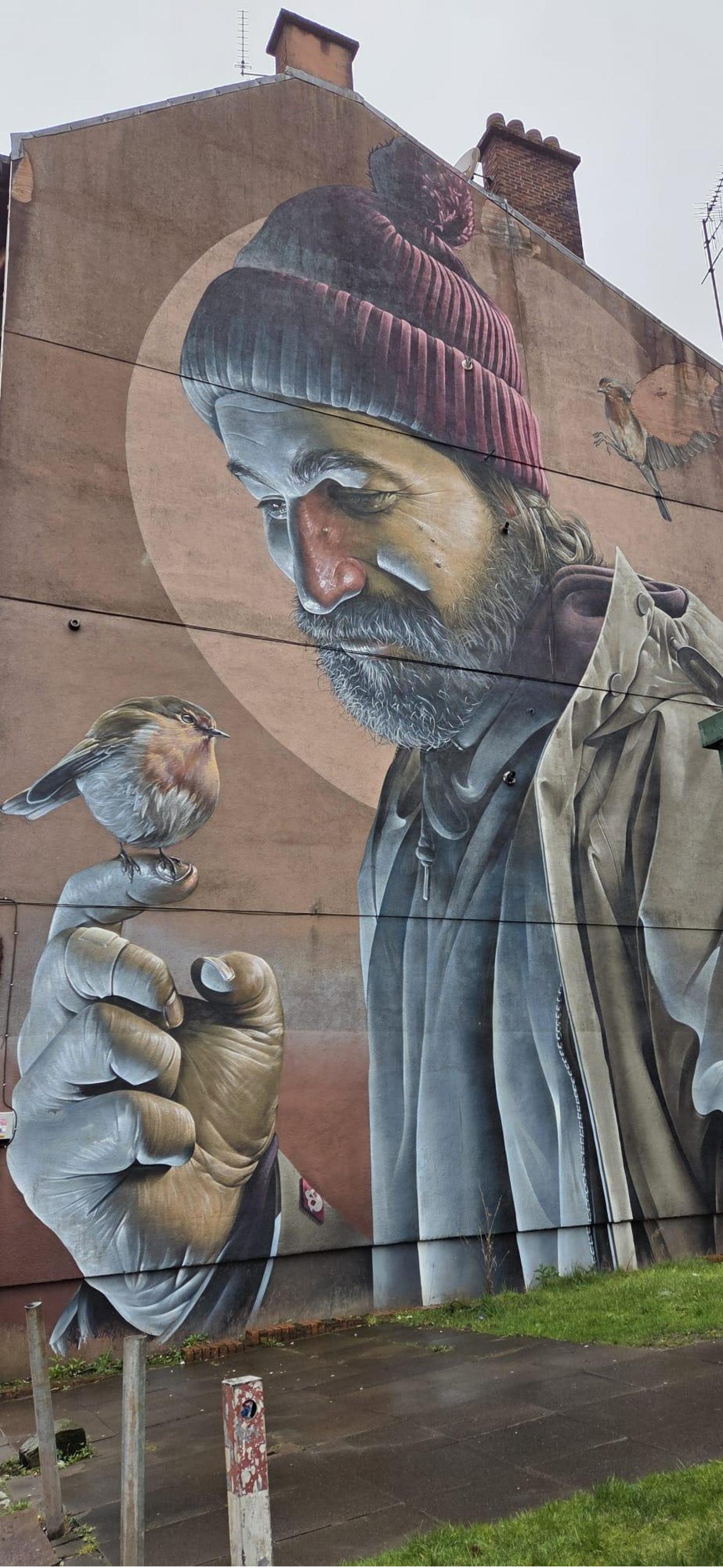 Saint Mungo. Otro de los trabajos de grandes dimensiones de Smug es el mural de San Mungo ataviado como un mendigo, muy cerca de la Catedral en la que reposan sus restos. No a mucha distancia, en la esquina High St y George St , el artista muestra al creador de Glasgow en brazos de su madre, St Enoch quien, al igual que el santo en su versión de adulto, tiene en su mano un petirrojo